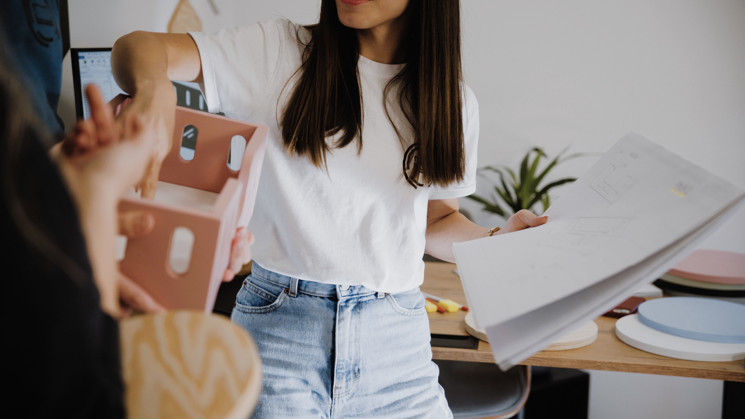 Kobieta w białej koszulce trzyma rysunki techniczne i rozmawia z drugą osobą pokazującą model różowego łóżka